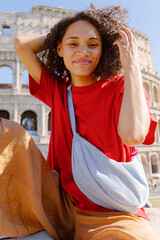 A young woman with vibrant curly hair, wearing a trendy red shirt and a stylish bag, poses joyfully in front of the iconic Colosseum in Rome, embodying the spirit of travel and adventure