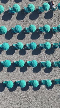 Vertical 4K top-down aerial video of turquoise umbrellas and lounge chairs arranged in perfect rows on Miami Beach, Florida, with morning sun casting long shadows, creating a vibrant geometric beach s