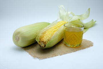 A clear glass filled with corn silk tea (Oksusu Cha), a traditional Korean herbal drink, is placed on a rustic fabric surface surrounded by fresh corn on the cob and corn husks. Ideal for wellness