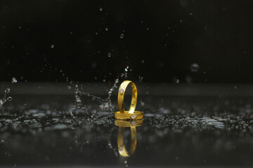 Two golden wedding bands reflecting on a wet surface with water splashes against a dark background,...