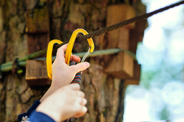 Yellow safety carabiner being attached by child to climbing rope, closeup of fastening process for outdoor zipline and amusement park activity ensuring security and protection. Selective focus
