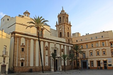 Obraz premium Full view of Santiago Apóstol Church and bell tower in Cádiz, Spain. Viewed from the plaza at sunset, with tall palm trees, baroque architectural details, stone facade, and warm atmosphere