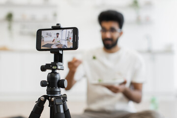 Young adult man sits in kitchen recording video about healthy eating habits. Scene shows him eating and using smartphone camera for vlog, promoting balanced diet and modern lifestyle.