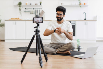 Caucasian adult man in casual outfit recording sports-related vlog discussing smart fitness devices. Room setting is a modern kitchen with exercise equipment visible.
