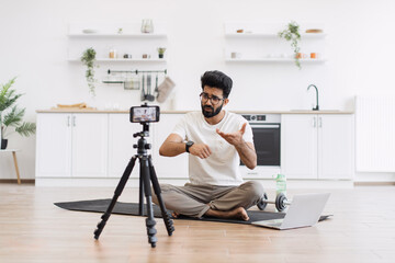 Caucasian adult man in casual outfit recording sports-related vlog discussing smart fitness devices. Room setting is a modern kitchen with exercise equipment visible.