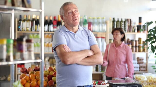 Senior wife stands behind pensioner husband and yells at companion. Angry man stand in front, he is sad and silent, ignores womans screaming. Couple find out their relationship near counter in store