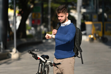 Young Caucasian man in a blue sweater checking his watch while standing with a bicycle, symbolizing...
