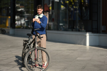 A Professional Caucasian man with backpack and bicycle stands outside, looking at his smartphone, blending fitness, technology, and urban life