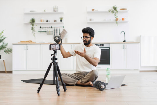Young man sits on yoga mat in kitchen filming workout tutorial with smartphone camera - Powered by Adobe
