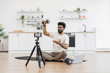 Adult man records fitness video blog at household kitchen, explaining dumbbell exercises. Sitting on floor with camera, tripod, and laptop, demonstrating healthy lifestyle ideas.