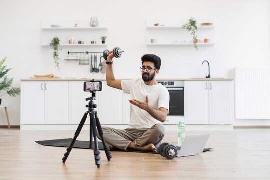 Adult man records fitness video blog at household kitchen, explaining dumbbell exercises. Sitting on floor with camera, tripod, and laptop, demonstrating healthy lifestyle ideas.