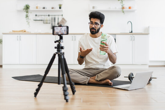 Young adult man recording fitness video in modern home discussing sports and hydration. Includes camera on tripod, yoga mat, water bottle, laptop, white rug, kettle, and minimalistic interior themes.