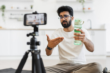 Young adult man with beard filming fitness blog discussing the value of hydration during workouts. Video setup includes smartphone on tripod indoors in a modern kitchen setting.