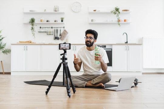 Young man with beard recording fitness video blog about hydration tips. Sitting in kitchen with water bottle mid-20s young man demonstrating healthy lifestyle online content creation.