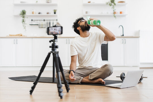 Young man in twenties seated in kitchen recording video about water importance for exercise. Scene includes mat, tripod camera, dumbbell, demonstrating creating fitness content in home environment.
