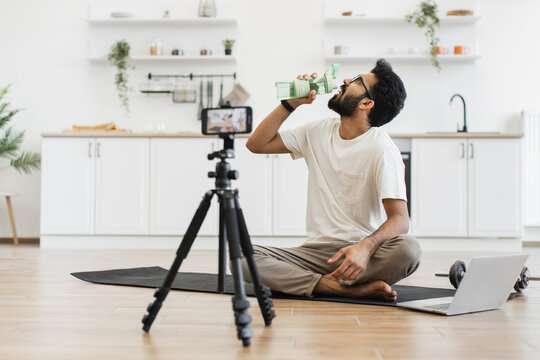 Young man in twenties seated in kitchen recording video about water importance for exercise. Scene includes mat, tripod camera, dumbbell, demonstrating creating fitness content in home environment.