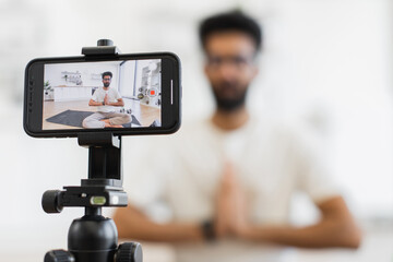 Young adult man meditating on yoga mat at home while recording his video blog focusing on fitness and wellness guidance using camera-mounted smartphone. Promotes calmness, mindfulness