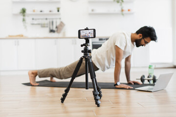 Young adult man in living room recording fitness blog video exercising on mat using laptop.