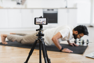Young adult man doing push-ups on exercise mat, recording fitness video blog using smartphone in indoor setting. Promotes healthy lifestyle and home workouts. Emphasizes dedication to fitness, health