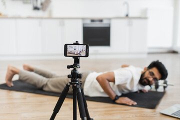 Young bearded man wearing casual clothes exercising on yoga mat at home recording fitness video while performing push-ups. Camera and laptop nearby show dedication to health and social media content.