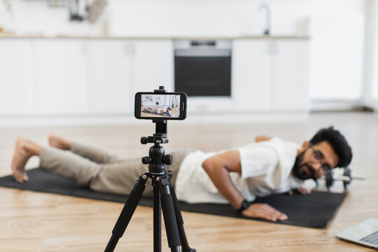 Young bearded man wearing casual clothes exercising on yoga mat at home recording fitness video while performing push-ups. Camera and laptop nearby show dedication to health and social media content.