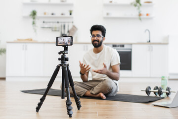 Adult man sitting on mat recording video about fitness, exercise benefits. Engaging vlog activity promoting health and workout. Explaining exercises suitable for environments during recording session.