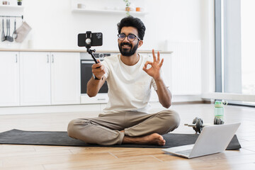 Young adult man with black beard and glasses records sports vlog at home. Sits on yoga mat in...
