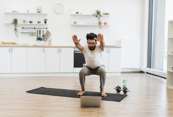 Adult male performing squat exercises on black mat in home kitchen setting using laptop.