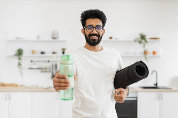 Young adult bearded man holding black yoga mat and green water bottle indoors in kitchen. Preparing for home fitness or yoga routine.