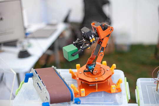 Robotic manipulator with orange arm on table. It holds a green cube and is part of a technology demonstration. Background includes blurred equipment. Basics of programming for children.