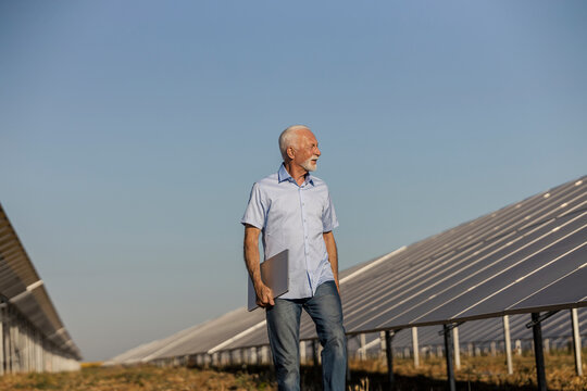 An old engineer walking by solar plant and holding laptop under armpit.