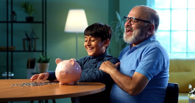 Indian grandfather teaching kid saving money in piggy bank at home, elderly man guiding grandson about financial discipline, inculcating good money habit for secure future, family bonding indoors
