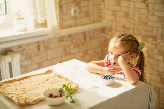 Little girl sits at the kitchen table with dough and berries, looking bored or thoughtful. Concept of childhood emotions, family life, homemade food, and cooking traditions.