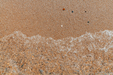 Close-up view of sea foam washing over golden sand on a beach. Transparent water and small pebbles create a natural coastal texture perfect for backgrounds and design elements.
