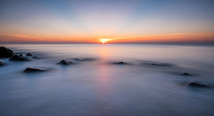Fototapeta premium Serene ocean sunset with soft, blurred waves gently washing over dark, wet rocks on the shore, creating a tranquil and peaceful atmosphere