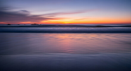 Obraz premium Serene ocean waves gently lap the shore during a breathtaking sunset, painting the sky with vibrant hues of orange, purple, and blue, creating a tranquil and picturesque coastal scene