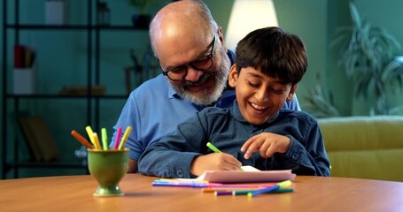 Indian grandfather helping kid drawing or coloring with pens, both sitting together at table in modern home, elderly man guiding grandson in creative activity, family bonding indoors happily - Powered by Adobe
