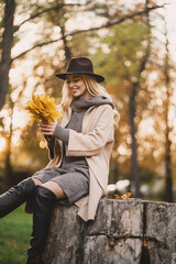 Happy young blonde woman on a sunny autumn day walking in the park with falling leaves in a stylish...