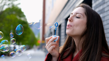 Young beautiful woman in burgundy coat blowing soap bubbles on city street. Freedom concept