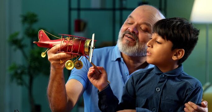 Indian grandfather helping kid coloring toy plane with brush and color, both sitting together at table in modern home, elderly man guiding grandson in creative artwork, family bonding moment