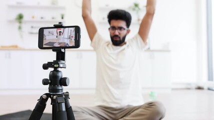 Male adult records fitness video blog using smartphone while using dumbbells for strength exercises. Seen seated in modern home interior, showing a health-focused and technology-driven lifestyle. - Powered by Adobe