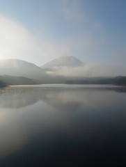 Obraz premium A serene landscape featuring a calm lake reflecting mountains shrouded in mist under a hazy sky