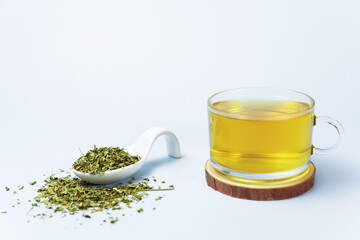 Moringa herbal tea in a glass cup with dried moringa leaves on a white spoon and a wooden coaster on a white background