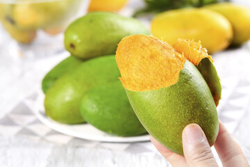 Hand holding peeled green mango revealing golden flesh with tropical fruits on white plate background