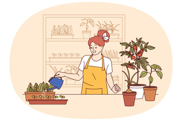 Woman florist grows house plants, uses watering can, standing near table and shelves with houseplants. Young girl florist enjoys own work, dressed in apron to avoid stains on clothes