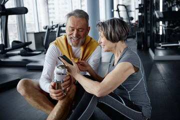 Mature couple are resting in a gym after exercises, looking at smartphote