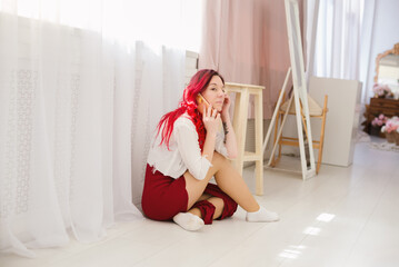 A young woman with vibrant red hair sits casually on a light wooden floor, engrossed in a phone...