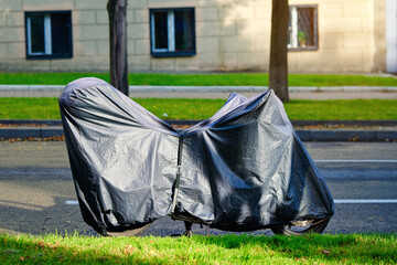 Parked motorcycle on roadside in city fully covered with black waterproof protective cover, uv resistance heat and wind protection with breathable anti moisture ventilation system, rain protection
