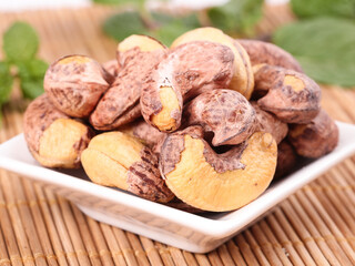 Roasted Cashews in White Bowl on Wooden Bamboo Mat with Green Leaves Background