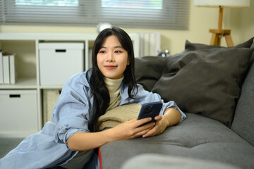 Obraz premium Young woman relaxing on sofa holding smartphone, smiling thoughtfully